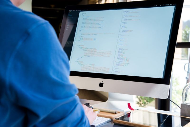 Homem de blusa azul usando um computador branco. Ao fundo vemos uma janela.